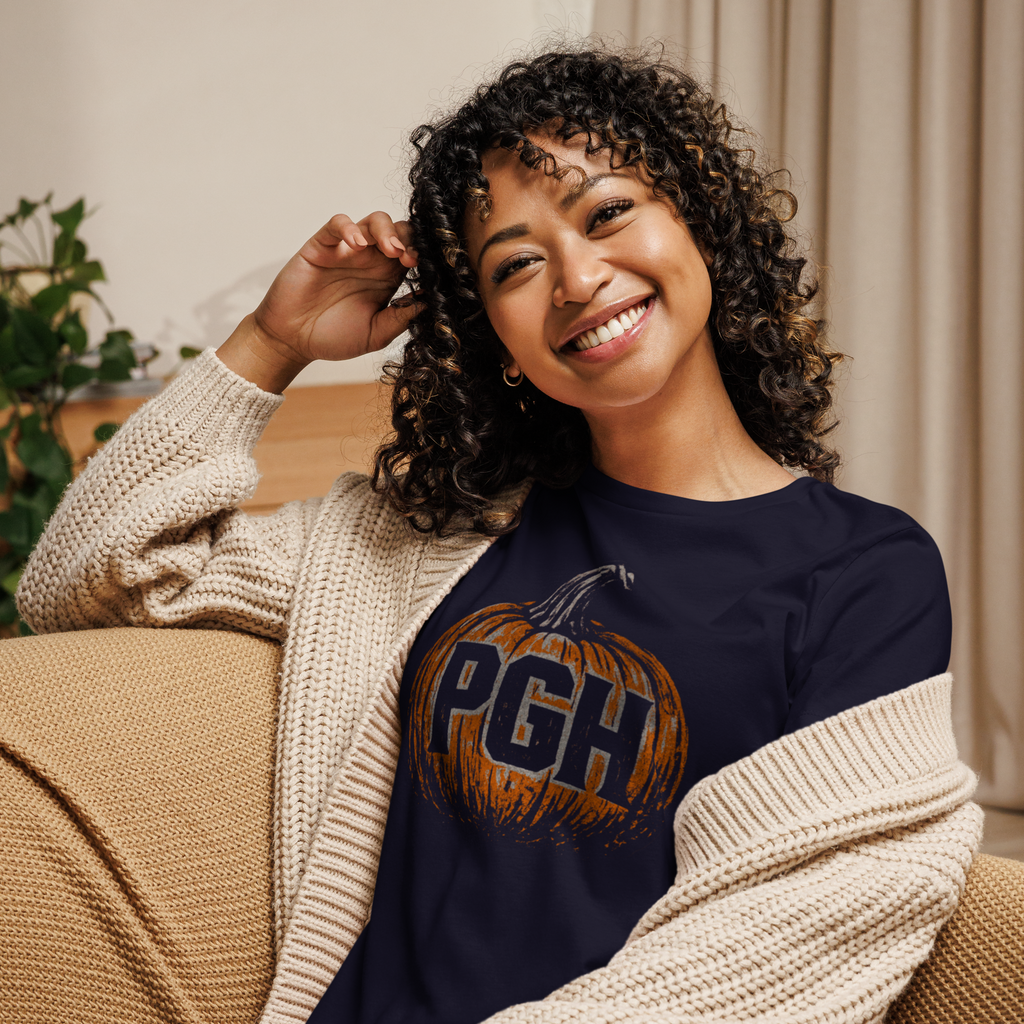Woman wearing a navy blue t-shirt with an orange pumpkin on it, with a cream colored sweater on, sitting on a couch.